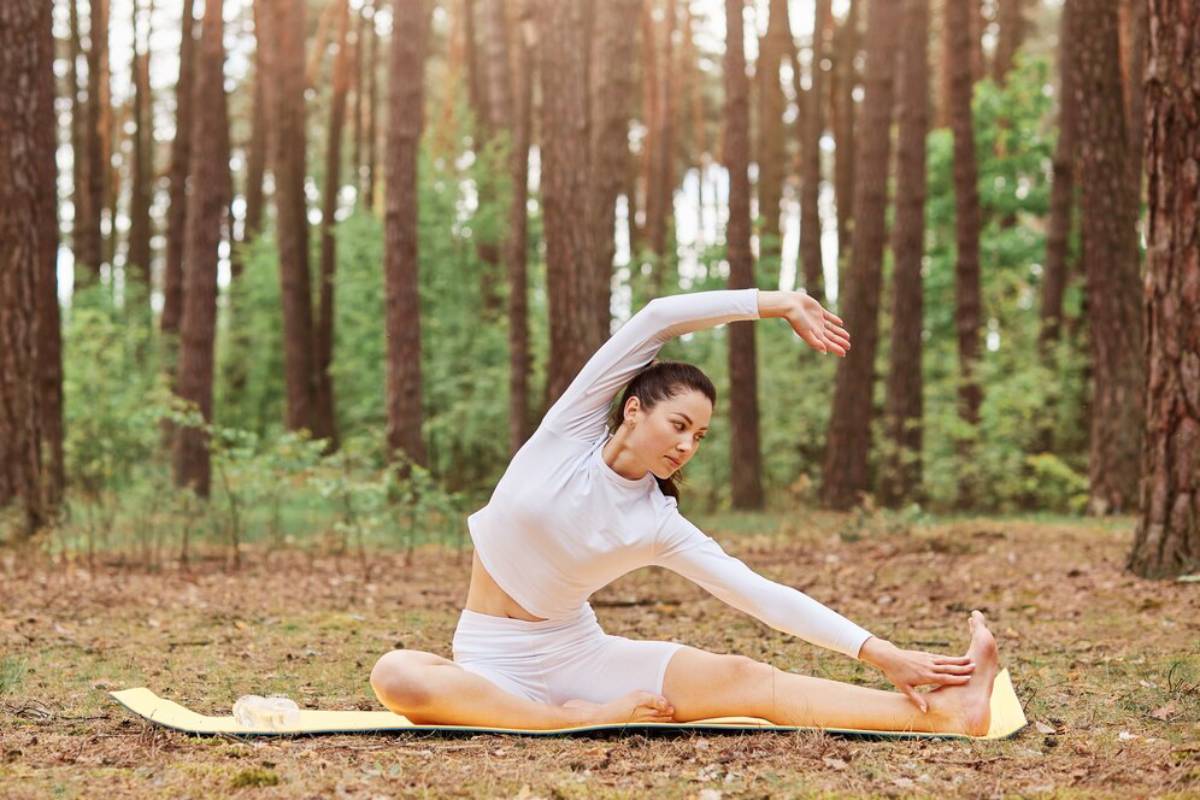 Woman performing a seated yoga stretch in a forest, wearing white athletic clothing.