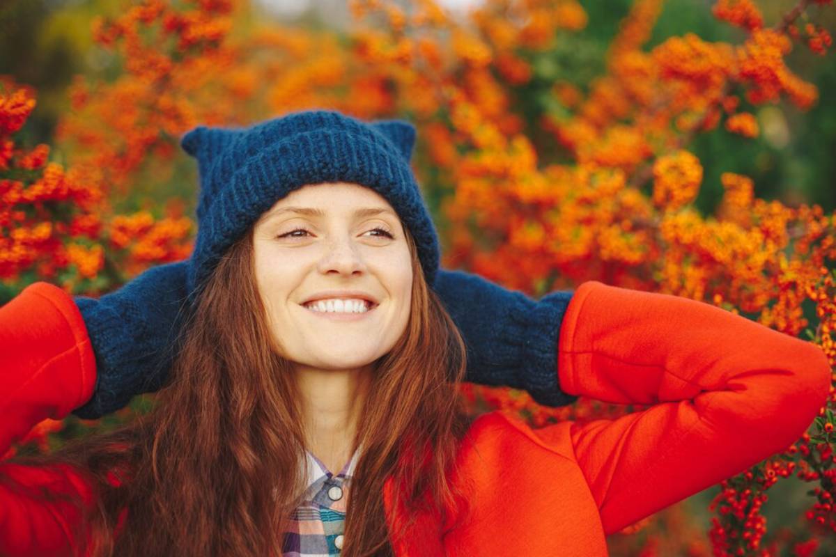 Smiling woman wearing a blue hat and red coat enjoying a vibrant autumn setting.