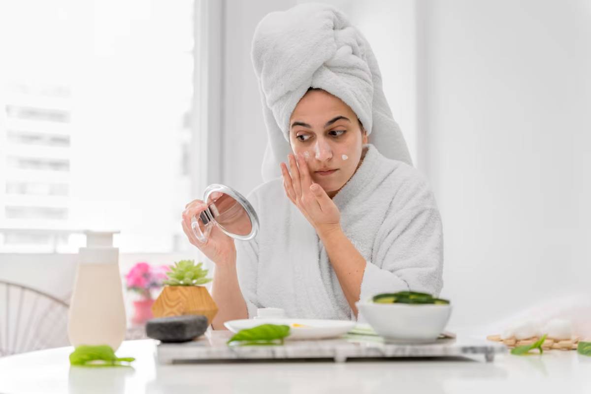 Woman in a bathrobe and towel on her head, applying facial cream while looking in a handheld mirror.