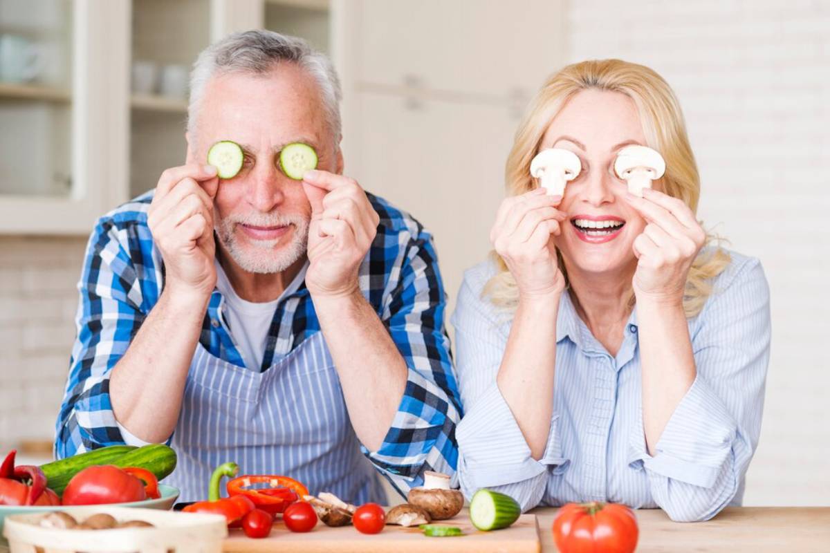Senior couple having fun with vegetables in a kitchen, holding slices of cucumber and mushrooms over their eyes, smiling.