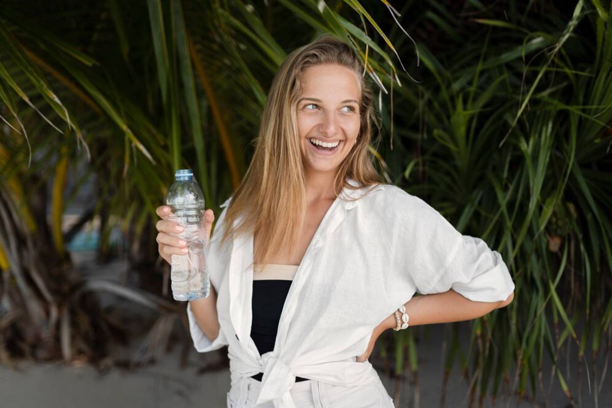A cheerful young woman holding a water bottle, standing among tropical plants, smiling and looking aside.