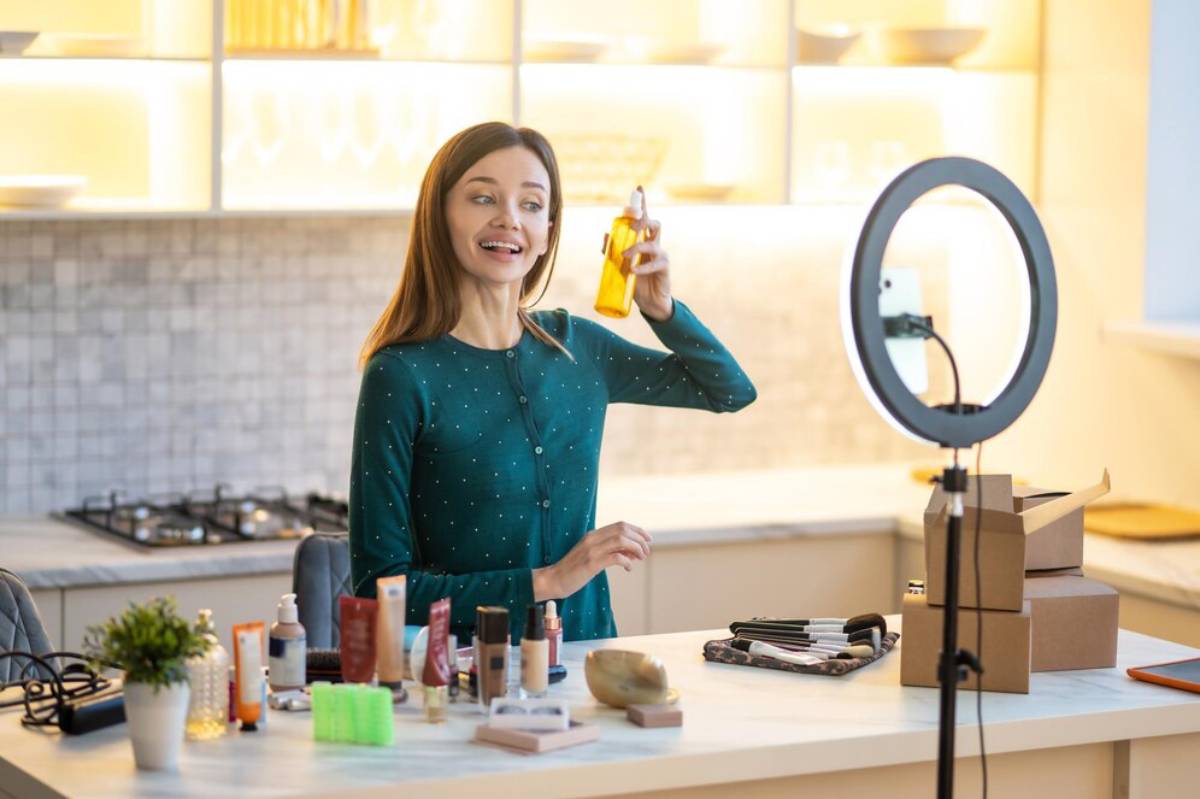 A woman in a teal cardigan holds a bottle in a bright kitchen, surrounded by various skincare and makeup products on the countertop.