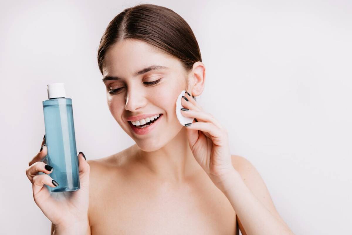 A woman smiles while holding a bottle of skincare product and using a beauty pad on her face.