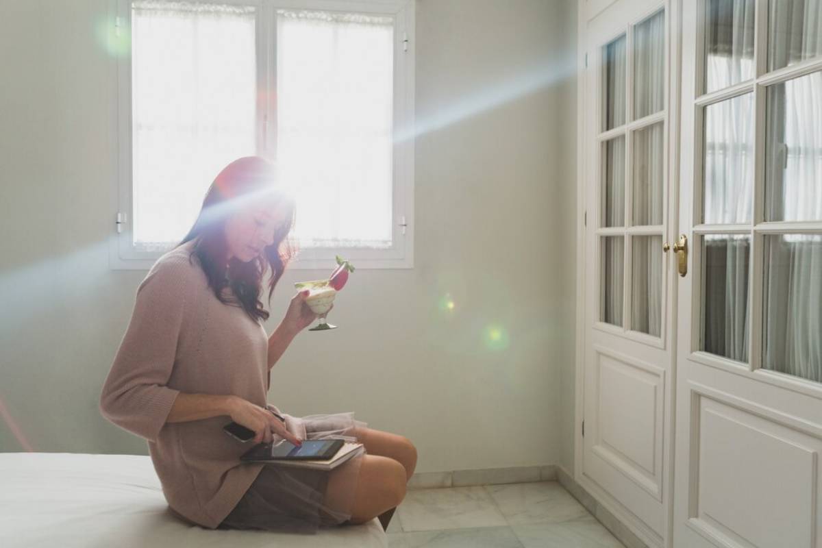 A person sits on a bed in a well-lit room, enjoying a drink and using a tablet, with a wardrobe and two windows in the background.