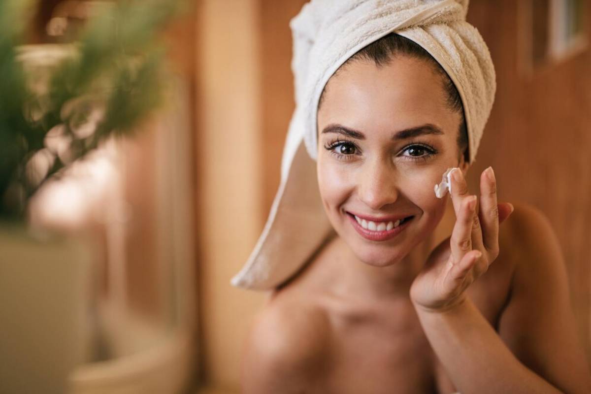 A smiling woman with a towel on her head applies cream to her face.