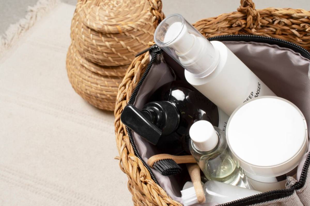 Open woven cosmetic bag with assorted skincare products and a straw hat on a neutral textile background.
