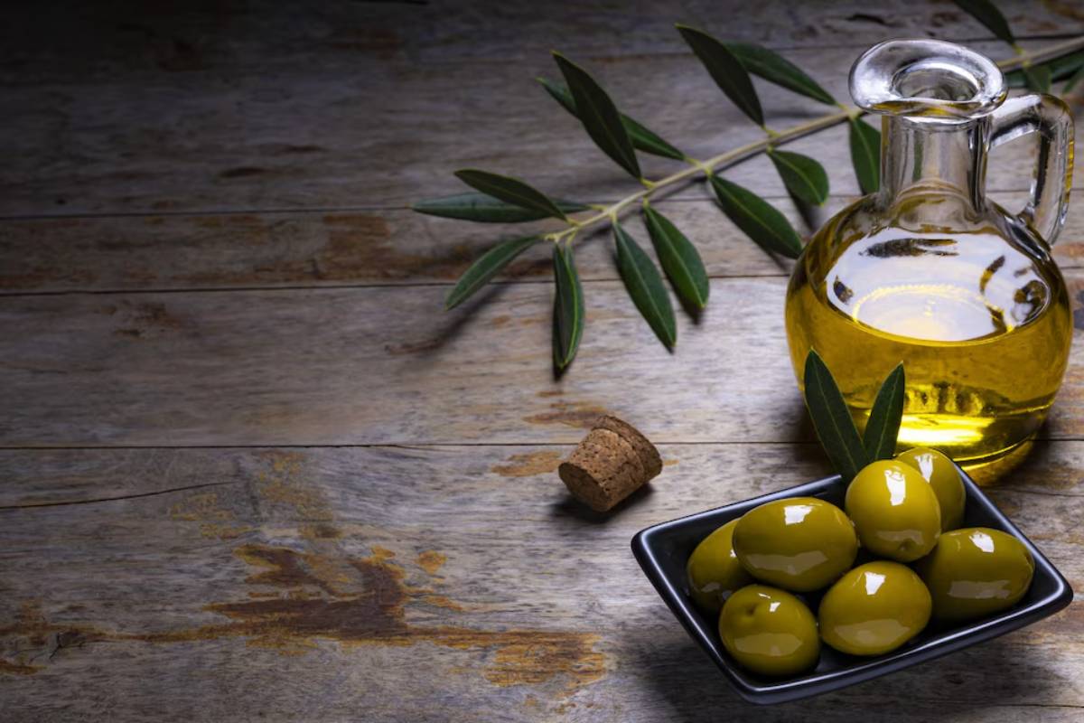 A glass jug of olive oil and a dish of green olives on a rustic wooden table.