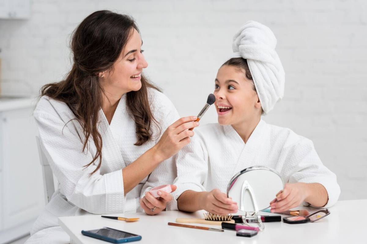 Two women in white robes engage in a beauty routine, surrounded by makeup products and a mirror on a bright, minimalist table.