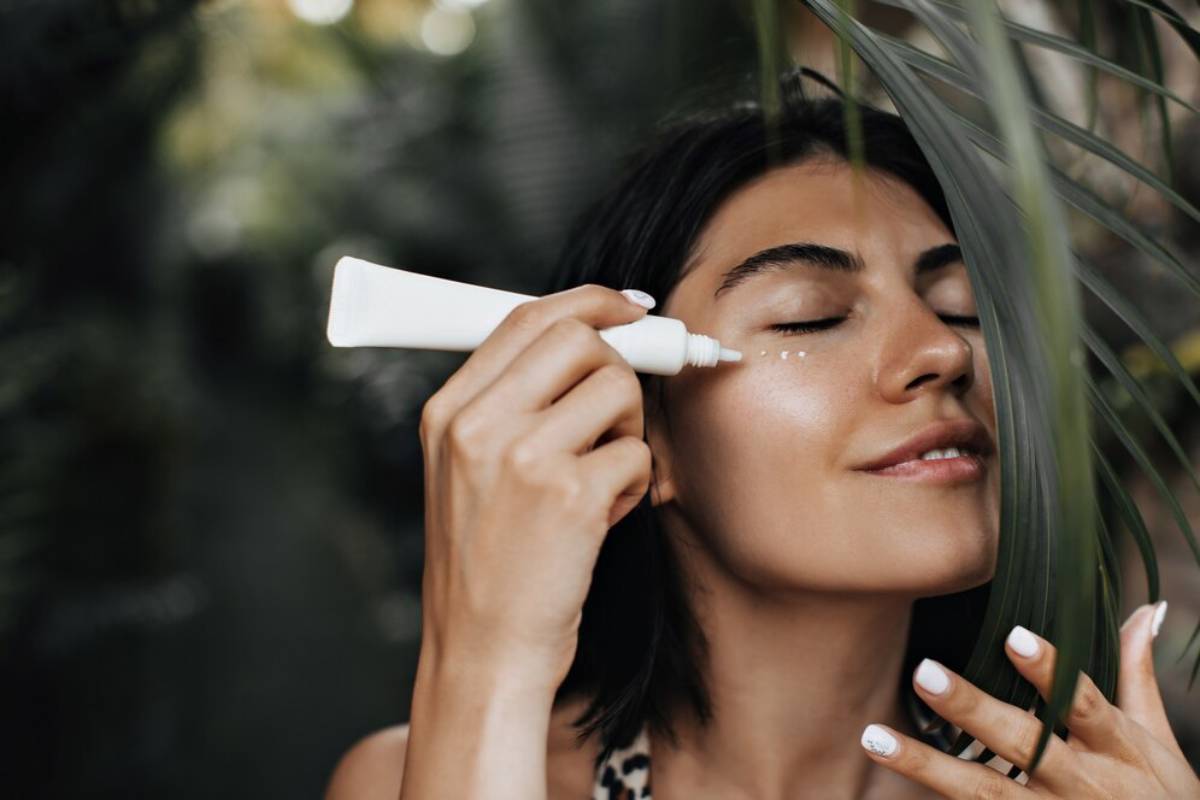 A woman applying serum to her face with a dropper bottle, surrounded by lush greenery.
