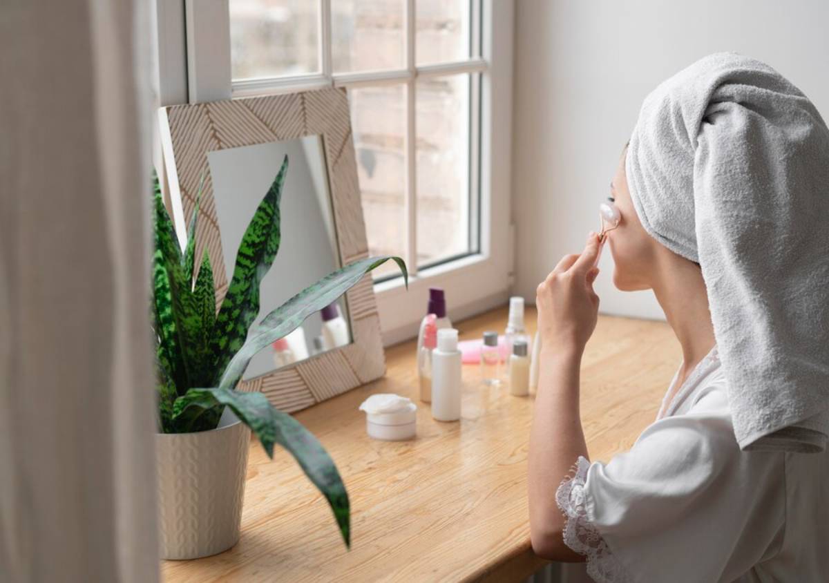 A woman with a towel on her head applies skincare in front of a mirror, surrounded by beauty products and a potted plant.