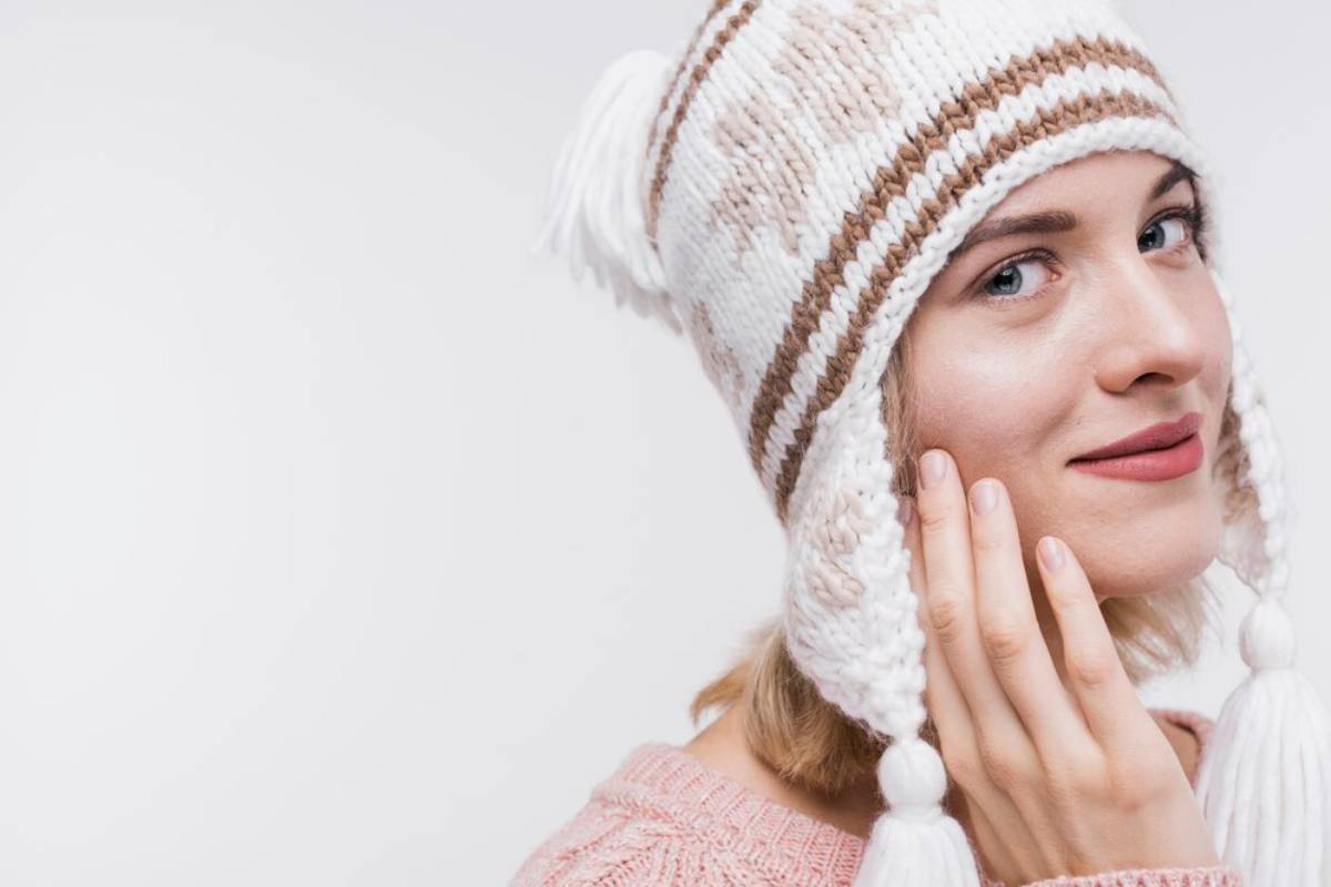 Young woman in a knitted white and brown hat smiling and touching her cheek against a light background.